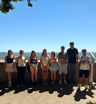 Eine Gruppe von sieben jungen Menschen steht auf einer Aussichtsplattform mit Blick über eine Stadtlandschaft bei sonnigem Wetter und blauem Himmel. Neben ihnen steht ein festinstalliertes Fernrohr.