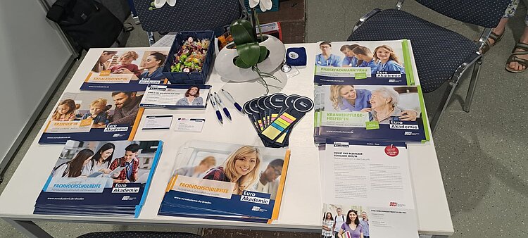 Informationsstand der Euro Akademie mit verschiedenen Flyern zu Ausbildungsangeboten wie Sozialassistentin, Erzieherin, Pflegefachmann/-frau, Fachhochschulreife und Krankenpflegehelfer*in. Auf dem Tisch befinden sich auch Werbeartikel wie Kugelschreiber, Haftnotizen, Visitenkarten, Bonbons und ein dekoratives Blumenarrangement.