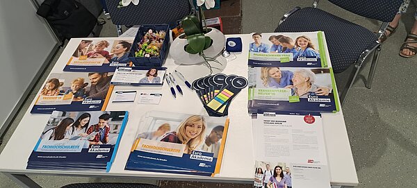 Informationsstand der Euro Akademie mit verschiedenen Flyern zu Ausbildungsangeboten wie Sozialassistentin, Erzieherin, Pflegefachmann/-frau, Fachhochschulreife und Krankenpflegehelfer*in. Auf dem Tisch befinden sich auch Werbeartikel wie Kugelschreiber, Haftnotizen, Visitenkarten, Bonbons und ein dekoratives Blumenarrangement.