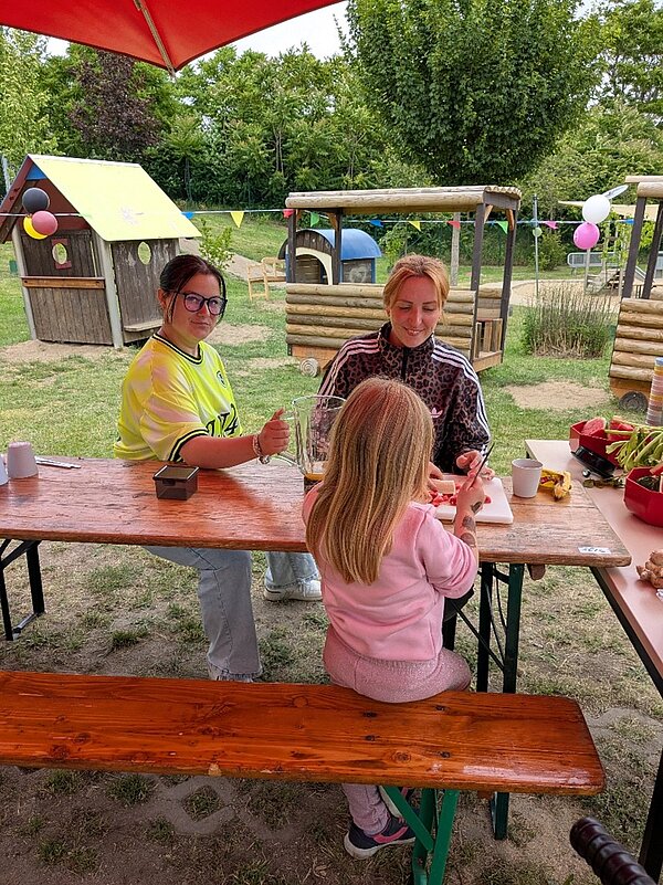 An einem Tisch im Außengelände bereiten Erwachsene gemeinsam mit einem Kind frisches Obst für eine Aktion vor.