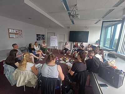 Schülerinnen und Schüler sowie Lehrkräfte sitzen im Klassenraum in einem Stuhlkreis, in der Mitte liegen bunte Moderationskarten.