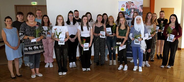Zu sehen ist eine große Gruppe von Schülerinnen und Schülern, die eine Blume und einen Flyer in der Han halten. Die Gruppe steht in einem Raum mit Parkettboden.