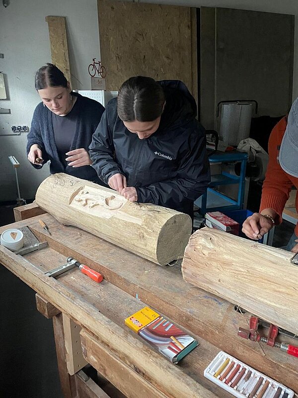 Zwei Personen arbeiten an einem großen Holzstamm auf einer Werkbank. Sie haben eine Eidechse herausgearbeitet. Im Hintergrund sind ein Regal und große Holzplatten zu sehen.  Auf der Werkbank liegen Werkzeuge und Kreide bereit.