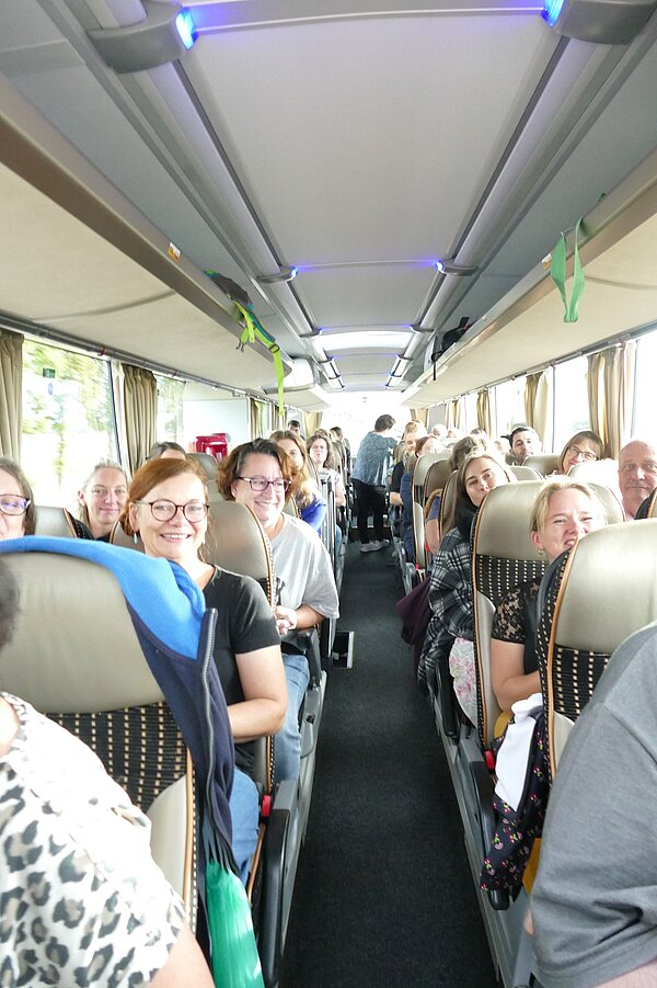 Gruppe von Erwachsenen sitzt während einer gemeinsamen Ausflugsfahrt in einem Reisebus und blickt teilweise lächelnd in die Kamera.