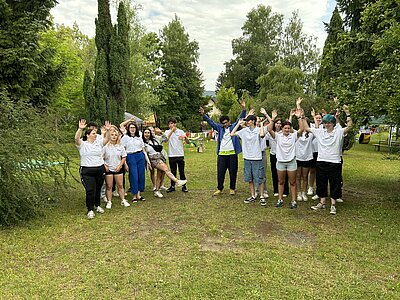 Gruppe junger Menschen steht auf einer Wiese und winkt lachend in die Kamera. Im Hintergrund Wimpelketten, Bäume und ein Teil des aufgebauten Spielbereichs.