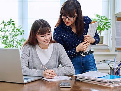 Zwei junge Frauen arbeiten gemeinsam an einem Schreibtisch: Eine sitzt mit Stift und Notizen neben einem Laptop, die andere steht daneben, hält Unterlagen in der Hand und unterstützt sie beim Lernen.