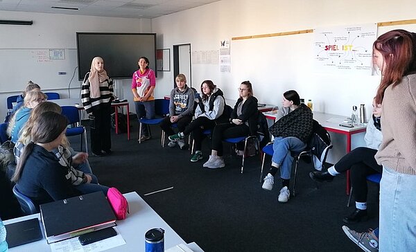 Sozialassistent*innen vertiefen Wissen über Cybermobbing Eine Gruppe von Schülern und Schülerinnen sitzt in einem Stuhlkreis in einem Klassenzimmer. Eine Lehrkraft steht vorne und moderiert die Diskussion. Neben ihr steht einer Schülerin mit Kopftuch. An der Wand hängt ein Plakat mit der Aufschrift "SPIEL IST", das verschiedene Aspekte von spielerischem Lernen zeigt.