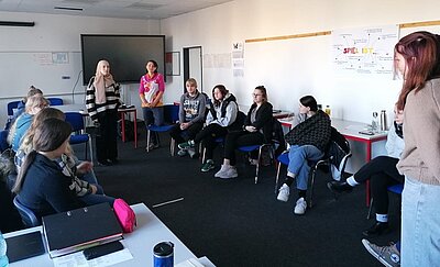  Eine Gruppe von Schülern und Schülerinnen sitzt in einem Stuhlkreis in einem Klassenzimmer. Eine Lehrkraft steht vorne und moderiert die Diskussion. Neben ihr steht einer Schülerin mit Kopftuch. An der Wand hängt ein Plakat mit der Aufschrift "SPIEL IST", das verschiedene Aspekte von spielerischem Lernen zeigt. 