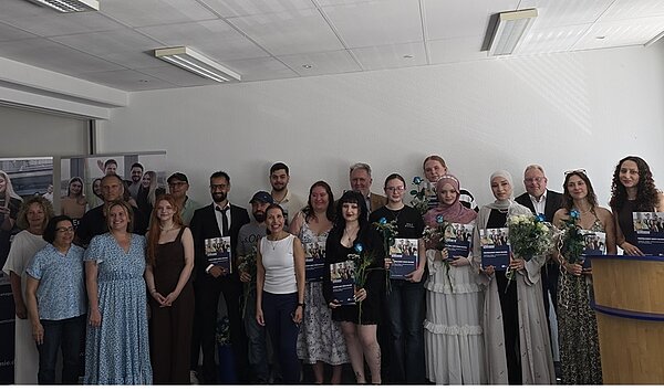 Die Gruppe der erfolgreichen Absolventinnen und Absolventen der Euro Akademie Hannover steht in einem hellen Raum nebeneinander und posiert für ein Erinnerungsfoto. Einige Personen halten große, rechteckige Urkunden oder Mappen in den Händen. Mehrere Personen tragen festliche oder elegante Kleidung, darunter Kleider und Anzüge. Einige halten Blumensträuße. Im Hintergrund sind weiße Wände und Deckenleuchten zu sehen. Links steht ein Roll-up mit Fotos von Menschen und Gebäuden. Rechts steht ein Rednerpult aus Holz.