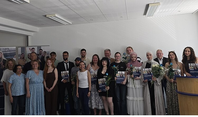 Die Gruppe der erfolgreichen Absolventinnen und Absolventen der Euro Akademie Hannover steht in einem hellen Raum nebeneinander und posiert für ein Erinnerungsfoto. Einige Personen halten große, rechteckige Urkunden oder Mappen in den Händen. Mehrere Personen tragen festliche oder elegante Kleidung, darunter Kleider und Anzüge. Einige halten Blumensträuße. Im Hintergrund sind weiße Wände und Deckenleuchten zu sehen. Links steht ein Roll-up mit Fotos von Menschen und Gebäuden. Rechts steht ein Rednerpult aus Holz.