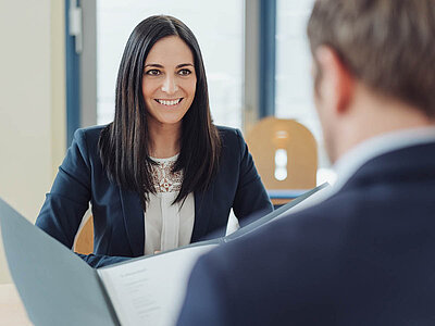 Auf dem Bild sind zwei Personen in einem professionellen Umfeld zu sehen. Eine Person hält ein geöffnetes Dokument, während die andere aufmerksam zuhört. Beide tragen formelle Kleidung, und der Raum ist hell mit Fenstern.