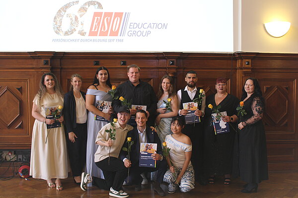 Gruppenbild mit festlich gekleideten Menschen. Die meisten Menschen halten Blumen und Zeugnismappen in den Händen. Im Hintergrund ist über einer holzvertäfelten Wand das 60-Jahre-Logo der ESO Education Group an die Wand projiziert. 