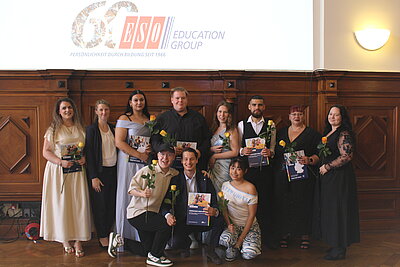 Gruppenbild mit festlich gekleideten Menschen. Die meisten Menschen halten Blumen und Zeugnismappen in den Händen. Im Hintergrund ist über einer holzvertäfelten Wand das 60-Jahre-Logo der ESO Education Group an die Wand projiziert. 