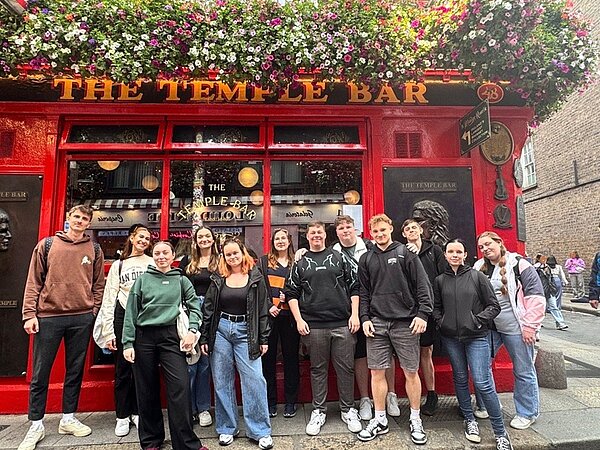 Gruppenfoto von Schülerinnen und Schülern der Euro Akademie Leipzig vor der berühmten roten Fassade der Temple Bar in Dublin. Die Jugendlichen stehen dicht beieinander, lächeln in die Kamera und tragen wetterangepasste Freizeitkleidung. Über ihnen hängt ein großes Blumenarrangement, und das goldene Logo ‚The Temple Bar‘ ist gut sichtbar. Das Bild entstand während eines kulturellen Ausflugs im Rahmen des Erasmus+ Aufenthalts in Irland.