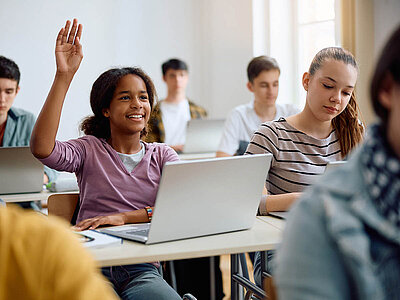 Eine Schülerin mit erhobener Hand sitzt an einem Tisch mit einem Laptop in einem Klassenzimmer, umgeben von anderen Schülern, die ebenfalls an Tischen sitzen.