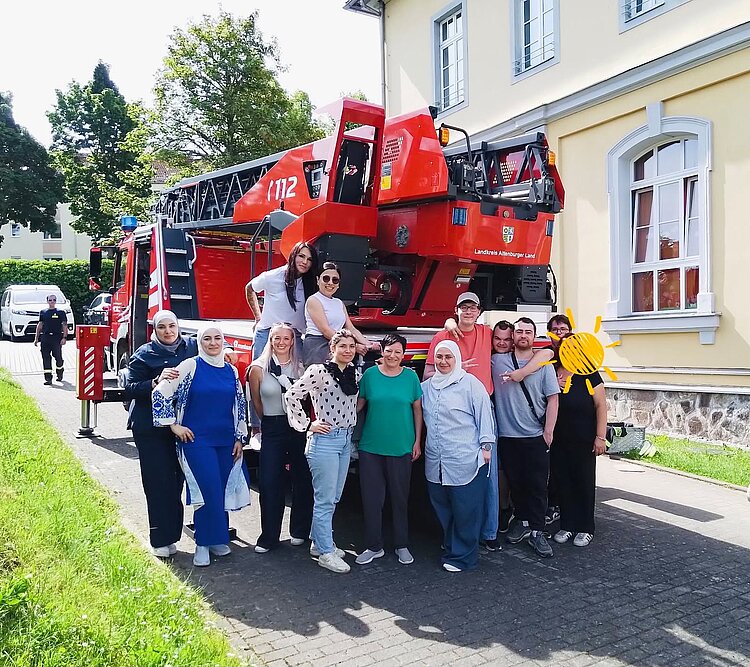 Eine Gruppe von Menschen steht lächelnd vor einem roten Feuerwehrfahrzeug mit der Aufschrift „Landkreis Altenburger Land“ und der Nummer „112“. Die Personen sind unterschiedlich gekleidet, einige tragen Kopftücher. Im Hintergrund sieht man ein gelbes Gebäude mit weißen Fensterrahmen und einen gepflasterten Weg.