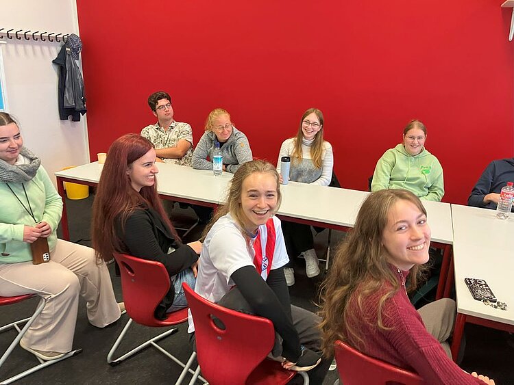 Wer sind die „Neuen“? – Kennenlernen der neuen Studierenden und Schüler*innen in Bamberg Eine Gruppe von neun jungen Erwachsenen sitzt lächelnd und entspannt in einem Seminarraum mit rot gestrichener Wand. Die Personen sind um zwei aneinandergestellte Tische herum arrangiert. Einige tragen Sweatshirts oder Pullover, andere haben Getränkeflaschen vor sich stehen. Die Stimmung wirkt locker und freundlich.