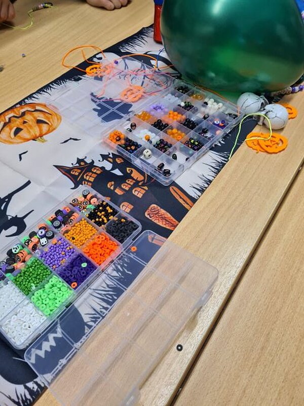 Auf einem Tisch liegt ein Plakat, auf dem typische Halloween-Symbole wie Kürbisse mit Gesichtern und Gespenster abgebildetet sind. Darauf liegen Boxen mit Bastelmaterial wie bunte Perlen und Schnüre und kleine Deko-Totenköpfe zu sehen. Im Hintergrund liegt ein dunkelgrüner Luftballon.