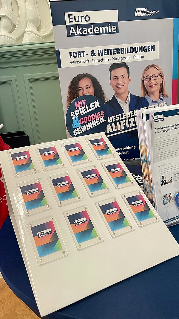 Ein weißes Präsentationsdisplay zeigt mehrere bunte Karten mit dem Logo der Euro Akademie. Im Hintergrund steht ein Roll-up mit Fotos von drei Personen, die freundlich lächeln. Darauf sind die Schriftzüge 'Euro Akademie', 'Fort- & Weiterbildungen', sowie weitere Begriffe zu sehen. Daneben befindet sich ein Prospektständer mit Informationsmaterial. Die Szene spielt in einem Innenraum mit grüner Wandvertäfelung.