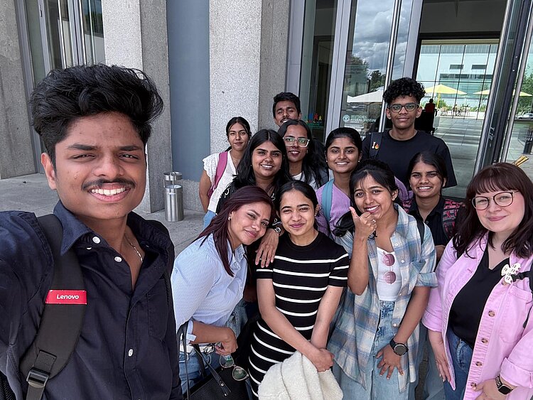 Eine Gruppe junger Menschen steht vor einem modernen Gebäude und lächelt in die Kamera. Die Gruppe wirkt fröhlich und freundlich, das Bild scheint ein Selfie zu sein, das bei einem Ausflug oder einer Exkursion aufgenommen wurde.