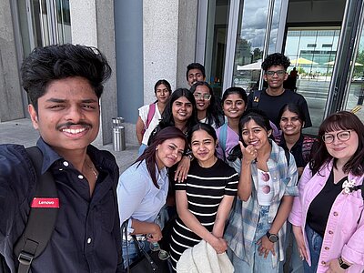 Eine Gruppe junger Menschen steht vor einem modernen Gebäude und lächelt in die Kamera. Die Gruppe wirkt fröhlich und freundlich, das Bild scheint ein Selfie zu sein, das bei einem Ausflug oder einer Exkursion aufgenommen wurde.