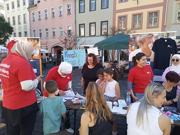 Menschen mit roten Euro Akademie T-Shirts betreuen Kinder und Erwachsene an einem Basteltisch auf einem Marktplatz, im Hintergrund bunte Häuserfassaden und ein Schild mit der Aufschrift „Kreativ-Insel“.