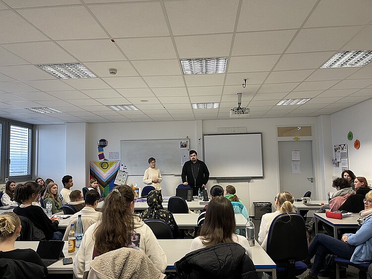 Besuch eines Bundestagsabgeordneten: Niklas Wagener an der Euro Akademie In einem Klassenzimmer stehen eine Lehrerin und der Bundestagsabgeordnete der Grünen, Niklas Wagener, vor der Tafel und hören einem*r Schüler*in zu, die*der gerade eine Frage stellt.