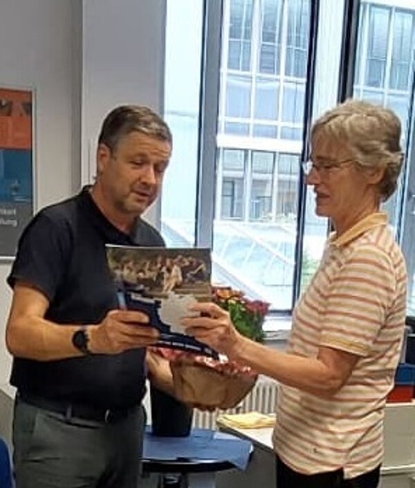 Ein Mann überreicht einer Frau einen Blumenstrauß und eine Mappe in einem Büro mit großen Fenstern.