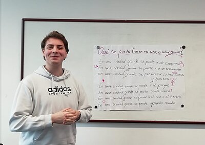 Ein Schüler steht lächelnd vor einem hangeschriebenen Plakat mit spanischen Sätzen.
