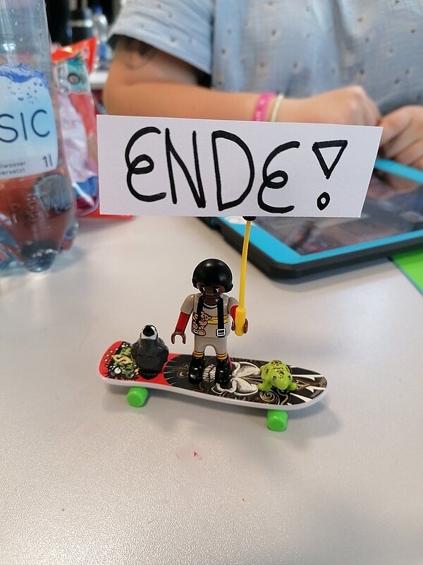 MeKoSax-Projekt „Trickfilmproduktion“ Eine schwarzhaarige Playmobilfigur steht auf einem Mini-Skateboard und hält ein Schild mit der Beschriftung Ende hoch.