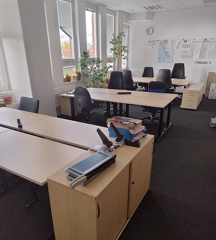 Blick in das neu gestaltete Lehrerzimmer, in dem nun höhenverstellbare Tische für ein ergonomisch gesundes Arbeiten stehen. An den Tischen stehen blau gepolsterte Bürostühle, an der Fensterfront zwei große Grünpflanzen, an der Wand hängt eine weiße Uhr und ein Kalender.