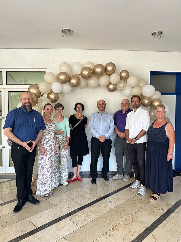 Unter einem Bogen aus weißen und goldenen Luftballons steht eine Gruppe von Lehrkräften und lächelt in die Kamera.