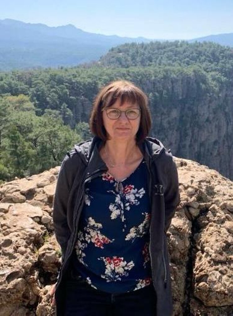 Eine dunkelhaarige Frau steht vor einem Felsen, hinter dem man eine Berglandschaft sieht.