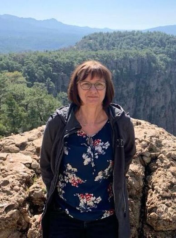 Eine dunkelhaarige Frau steht vor einem Felsen, hinter dem man eine Berglandschaft sieht.
