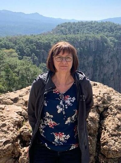Eine dunkelhaarige Frau steht vor einem Felsen, hinter dem man eine Berglandschaft sieht.