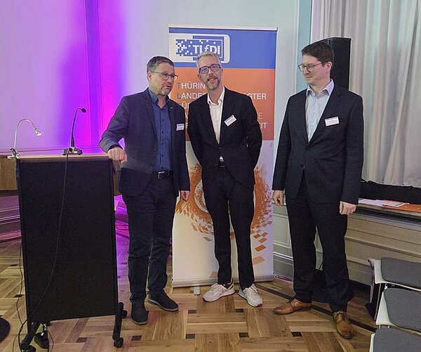 Leiter der Euro Akademie Erfurt und Jena, Uwe Schöneck, steht mit beiden Referenten an einem Rednerpult und unterhält sich. Alle tragen dunkle Anzüge und eine Brille.