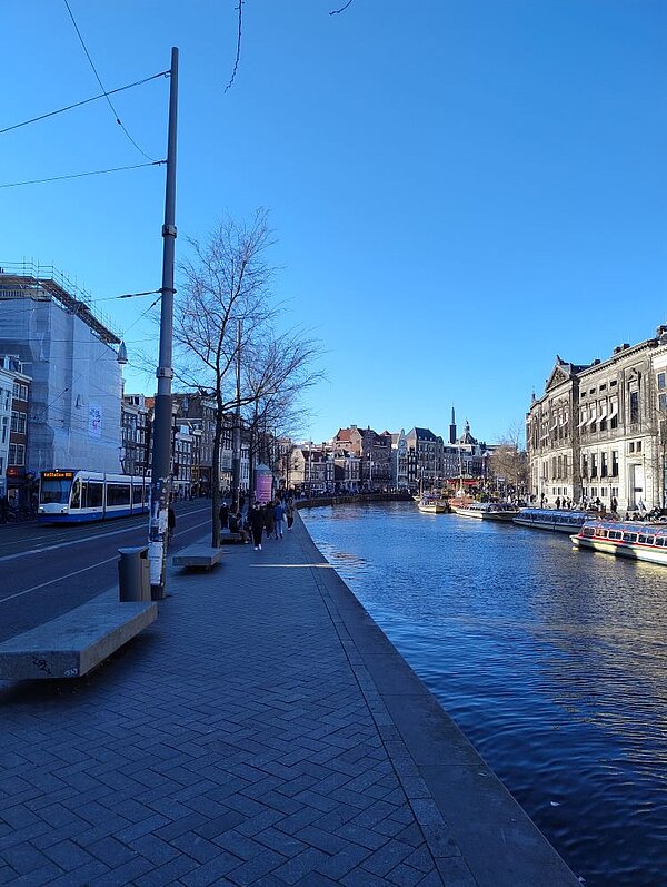 Ein Kanal in Amsterdam, gesäumt von historischen Gebäuden und Bäumen. Ganz links fährt eine Straßenbahn. Die Sonne scheint, und der Himmel ist blau. Im Hintergrund sind weitere Häuser  zu erkennen.