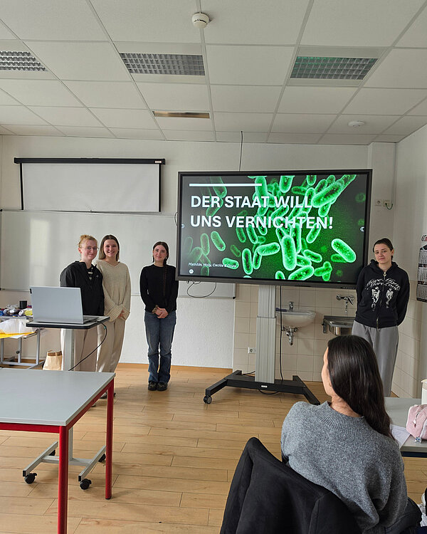 Drei Personen stehen vor einem Bildschirm, auf dem der Titel „Der Staat will uns vernichten!“ von Mathilde, Holly, Cecilla und Ömer zu sehen ist. Die Folie zeigt ein Bild von Bakterien.