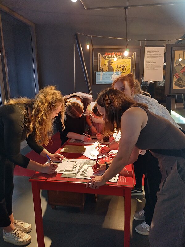 Eine Gruppe junger Frauen arbeitet konzentriert an einem roten Tisch, füllt Arbeitsblätter aus und schreibt mit Stiften. Im Hintergrund eine Ausstellung mit historischen Plakaten und Infotafeln.
