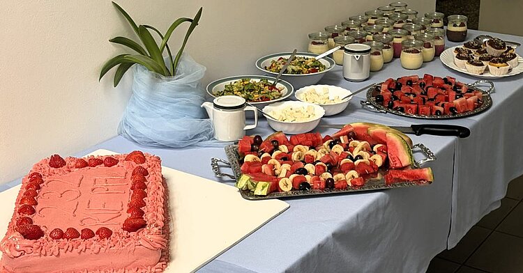 Ein weiterer Buffettisch mit süßen Speisen. Im Vordergrund steht eine große, mit Erdbeeren verzierte Torte. Daneben gibt es Obstplatten mit Wassermelone, Bananen und Trauben, kleine Desserts in Gläsern, Cupcakes sowie Schüsseln mit Obstsalat und Käse. Eine Topfpflanze und ein Milchkännchen ergänzen das Gedeck.