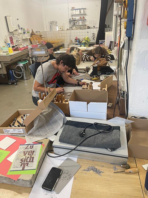 In einem hellen Atelier arbeiten mehrere Personen konzentriert an ihren Arbeitsplätzen. Im Vordergrund liegt ein Tisch mit Materialien wie einer Metallplatte, bunten Papieren und Werkzeug.