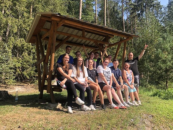 Die Auszubildenden zu Pflegefachkräften posieren bei Sommerwetter in einem offenen Waldhäuschen für ein Gruppenbild.