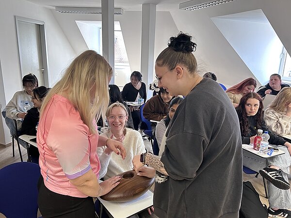 In einem hellen Seminarraum mit Dachschrägen stehen zwei Frauen im Vordergrund an einem Tisch. Die Frau links trägt ein rosa Oberteil, die Frau rechts ein dunkles Sweatshirt. Beide beugen sich über eine Person, die auf einem Stuhl sitzt. Im Hintergrund sitzen mehrere Personen an Tischen, einige schreiben oder schauen nach vorn. Auf den Tischen liegen Hefte, Stifte und Getränkeflaschen. Die Atmosphäre wirkt konzentriert.