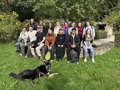 Die Auszubildenden posieren, gemeinsam mit Schäferhund-Mix Bailey, gut gelaunt auf einer Steinmauer sitzend für ein Gruppenfoto.