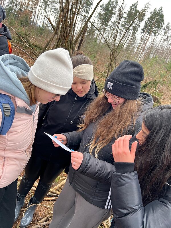 Vier Schülerinnen lesen einen Zettel, den eine von ihnen in der Hand hält. Sie befinden sich in einem Wald.