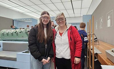 Zwei junge Frauen stehen in einem Ausstellungsraum nebeneinander, haben Schutzbrillen auf und lächeln.