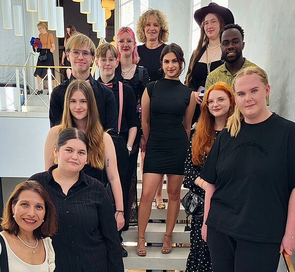 All That Jazz! Englischunterricht im Opernhaus Hannover Eine Gruppe von Menschen steht auf einer Treppe in einem modernen Gebäude. Alle tragen festliche oder elegante Kleidung. Im Hintergrund sind moderne Deckenleuchten sichtbar.