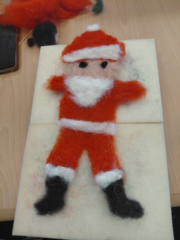 Gefilzte Weihnachtsmannfigur in Rot mit weißem Bart und Mütze liegt auf einer rechteckigen Filzunterlage auf einem Tisch.