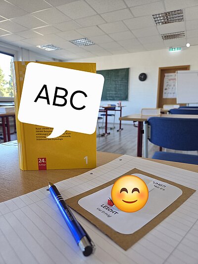 In einem Klassenzimmer steht in Nahaufnahme ein Wörterbuch auf dem Tisch. Davor liegt ein Stift und kleiner Schokoriegel.