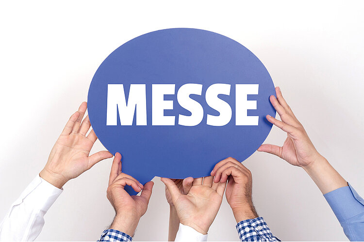 Mehrere Hände halten ein blaues Schild mit der Aufschrift "Messe" in weißen Lettern hoch. 